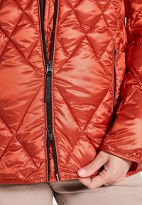 Gequiltede oranje pufferjack met een rits aan de voorkant, zwarte hardware en een gladde, glanzende textuur. Close-up van het taillegebied en de hand.