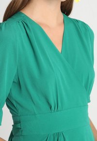 Vestido verde de wrap con mangas cortas abullonadas, que presenta detalles de fruncido en los hombros y la cintura, hecho de tela ligera.