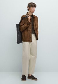 Brown suede jacket with a button front, beige wide trousers, dark sweater, and brown shoes. Carrying a black tote bag.