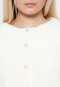 Vit ribbad cardigan i mjukt material, med rund halsringning och pärlknappar längs framsidan.