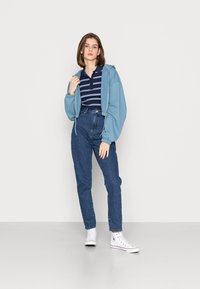 Hoodie azul claro com fecho de correr, polo listrado em azul-marinho, jeans azuis de cintura alta e ténis brancos. Conjunto casual com um corte descontraído.
