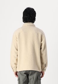 Beige Fleecejacke mit strukturiertem Finish, Kragen und geknöpften Ärmelbündchen, kombiniert mit olivgrünen Cargo-Hosen.