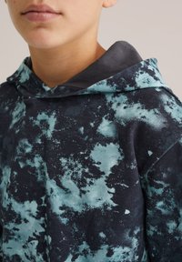 Felpa con cappuccio caratterizzata da una base blu scuro con motivi di schizzi turchesi, realizzata in tessuto morbido, che presenta un fitting rilassato e una superficie testurizzata.