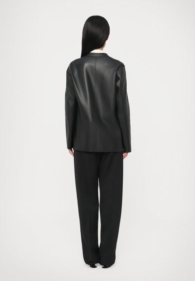 Melnas ādas žakete ar taisnu griezumu, minimālu detaļām un izgrieztu apkakli, kopā ar vaļīgām melnām biksēm.