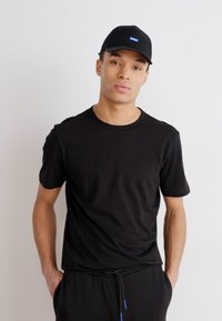 T-shirt nera a maniche corte realizzata in tessuto morbido, abbinata a un cappellino nero con un logo rettangolare blu, indossata in modo casual con pantaloni scuri.