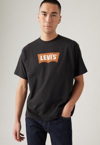 Čierne bavlnené tričko s oranžovým logom Levi's na prednej strane, s okrúhlym výstrihom, krátkymi rukávmi a voľným strihom. Nosené s tmavými džínsami.