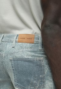 Svijetloplave traperice s teksturiranim uzorkom, s smeđim kožnim zakrpom na stražnjoj strani pojasa s natpisom "SAMSOE SAMSOE".