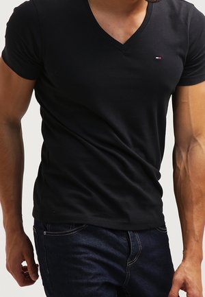 Mand iført sort V-hals T-shirt og mørkeblå jeans, med muskuløse arme synlige, stående mod en ensfarvet baggrund.