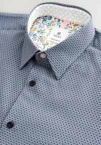 Cămașă albastră cu un model, realizată dintr-un material texturat, având un imprimeu floral pe interiorul gulerului. Două butoane din plastic negru și roz. Eticheta de marcă este vizibilă.