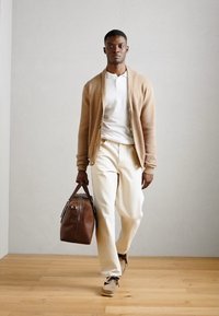 Tan pleten cardigan, bela polo, kremne hlače, drži rjavo usnjeno torbo. Lesena tla, nevtralna stenska ozadja. Preprost, modern dizajn.