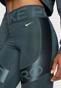 Tamnozeleni tajice s visokim pojasom na kojem piše "NIKE PRO" te mala bijela Nike oznaka. Glatka tkanina s kontrastnim sjajnim detaljima.