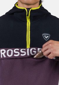 Pullover in fleece, blu navy e viola. Include una chiusura con zip frontale con accenti giallo fluo, una patch con il logo e una tasca nascosta per riporre oggetti.