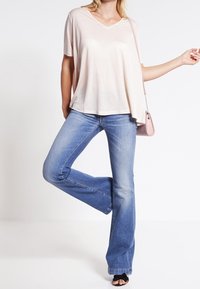 Kvinna i en ljusrosa lös V-ringad topp, blå utsvängda jeans, svarta öppna klackar och en rosa axelväska, som står med ett ben upphöjt.