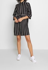 Abito camicia a righe bianco e nero con maniche lunghe, chiusura a bottoni sul davanti e due tasche sul petto, stretto in vita da una cintura.