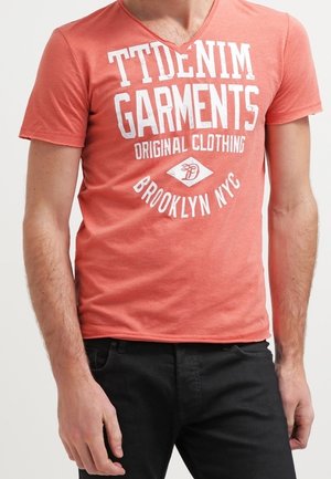 Mann trägt ein korallenrotes V-Ausschnitt-T-Shirt mit weißem Text "TTDENIM GARMENTS ORIGINAL CLOTHING BROOKLYN NYC" und schwarze Hose.