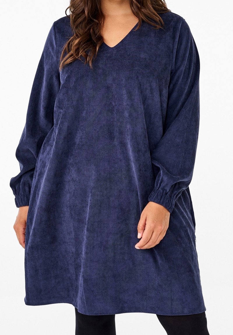 Robe en velours côtelé bleu marine avec un col en V. Elle présente des manches longues avec des poignets élastiques et une silhouette décontractée. Texture douce et longueur mi-cuisse.
