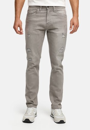 Mand iført lysegrå slidte slim-fit jeans og hvide sneakers, stående med hænderne afslappet langs siden.