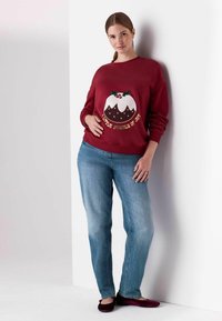 Rode sweater met een feestelijk ontwerp van een witte pudding, hulstaccenten en gouden tekst. Gecombineerd met lichtblauwe jeans en bordeauxkleurige flats.