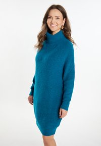 Rochie tricotată de culoare teal, cu guler înalt, ribotat, manșoni lungi și o croială lejeră. Textura este moale și ușor pufoasă.