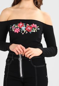 Schwarzes Off-Shoulder-Oberteil mit floralen Stickereien in Pink, Rot und Blau. Lange Ärmel, taillierte Passform und weicher, dehnbarer Stoff.
