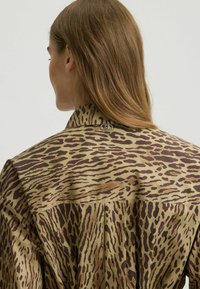 Bruine en beige shirt met een luipaardprintpatroon, voorzien van een puntige kraag en een metalen logoaccent op de achterkant. Glad textuur van de stof.