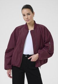 Veste bombardier bordeaux avec fermeture éclair à l'avant, col côtelé et coupe surdimensionnée. Elle est dotée de poches latérales et d'une texture lisse et brillante.