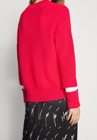 Pull en maille côtelée rouge avec des manches longues et des accents de poignets blancs, présentant une coupe décontractée. Porté par-dessus une jupe noire à motifs.