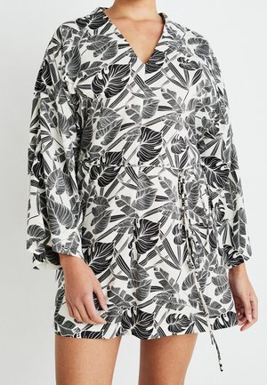 Femme portant une robe à motif de feuilles tropicales noir et blanc avec des manches longues amples et une ceinture à la taille, debout devant un fond uni.