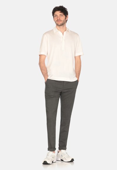 Polo shirt beige chiaro con colletto e maniche corte, abbinato a pantaloni grigio scuro e sneakers bianche, in piedi su uno sfondo neutro.