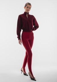 Burgundowa skórzana bluza z krótkim rękawem, z wysokim kołnierzem i zamkiem, zestawiona z dopasowanymi legginsami z bocznymi rozcięciami oraz czarnymi, spiczastymi butami.