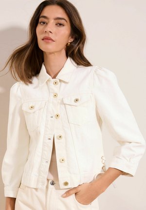 Donna con capelli castani fino alle spalle che indossa una giacca bianca con bottoni e spalle imbottite, mano in tasca, sfondo neutro.