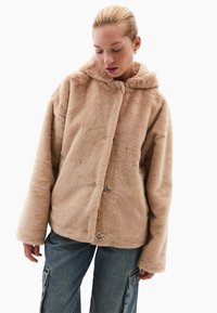 Beige umělý kožich s kapucí, se soft dotekem, volným střihem a dvěma stříbrnými patentkami. Nosený přes džíny.