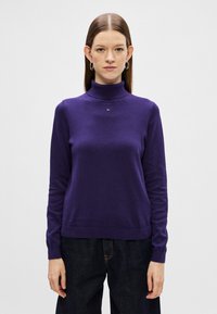 Fioletowy sweter z golfem wykonany z miękkiego materiału dzianinowego, z długimi rękawami i małym detalem logo przy wykończeniu dekoltu. W parze z ciemnymi dżinsami.