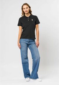 Zwarte polo met korte mouwen, drie knopen bij de hals en geborduurd logo. Gecombineerd met lichtblauwe uitlopende jeans en witte sneakers.
