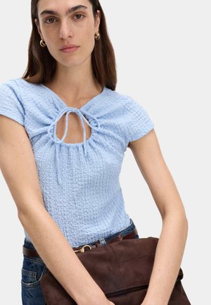 Vrouw draagt een lichtblauw gestructureerd topje met korte mouwen en een strikdetail aan de voorkant, blauwe jeans, gouden oorbellen in ringvorm en houdt een bruine leren clutch vast.