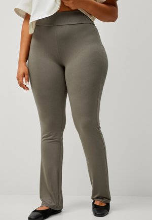 Rond persoon in olijfgroene hooggetailleerde legging en zwarte ballerina's, met een witte top tegen een effen achtergrond omhoog houdend.