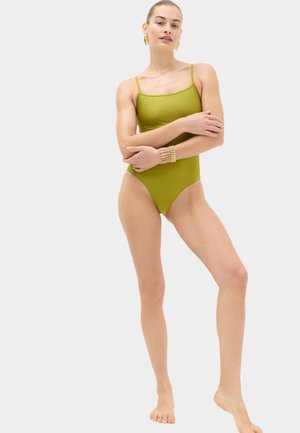 Femme debout pieds nus portant un maillot de bain une pièce vert olive avec des fines bretelles et des bracelets en or au poignet gauche.