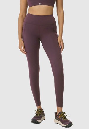 Vrouw draagt paarse leggings met hoge taille en zijzakken en een bijpassende sportbeha, gecombineerd met paarse en beige sportschoenen.