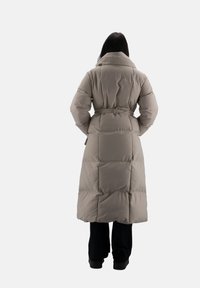 Lang beige pufferjakke med bælte i taljen, høj krave og kviltet tekstur. Vist fra ryggen med sorte bukser og støvler.