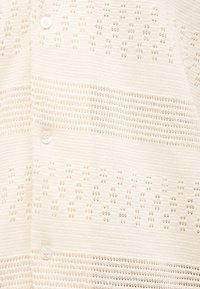 Κρεμ πλεκτή ζακέτα με ανάγλυφες οριζόντιες ρίγες και σχέδια ανοιχτής πλέξης. Περιλαμβάνει ένα στρογγυλό, διάφανο κουμπί κλεισίματος.