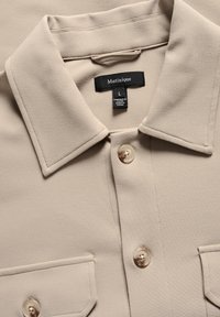 Camisa beige de botonadura con cuello, dos bolsillos frontales y una textura suave. Presenta una etiqueta de marca negra y botones transparentes.