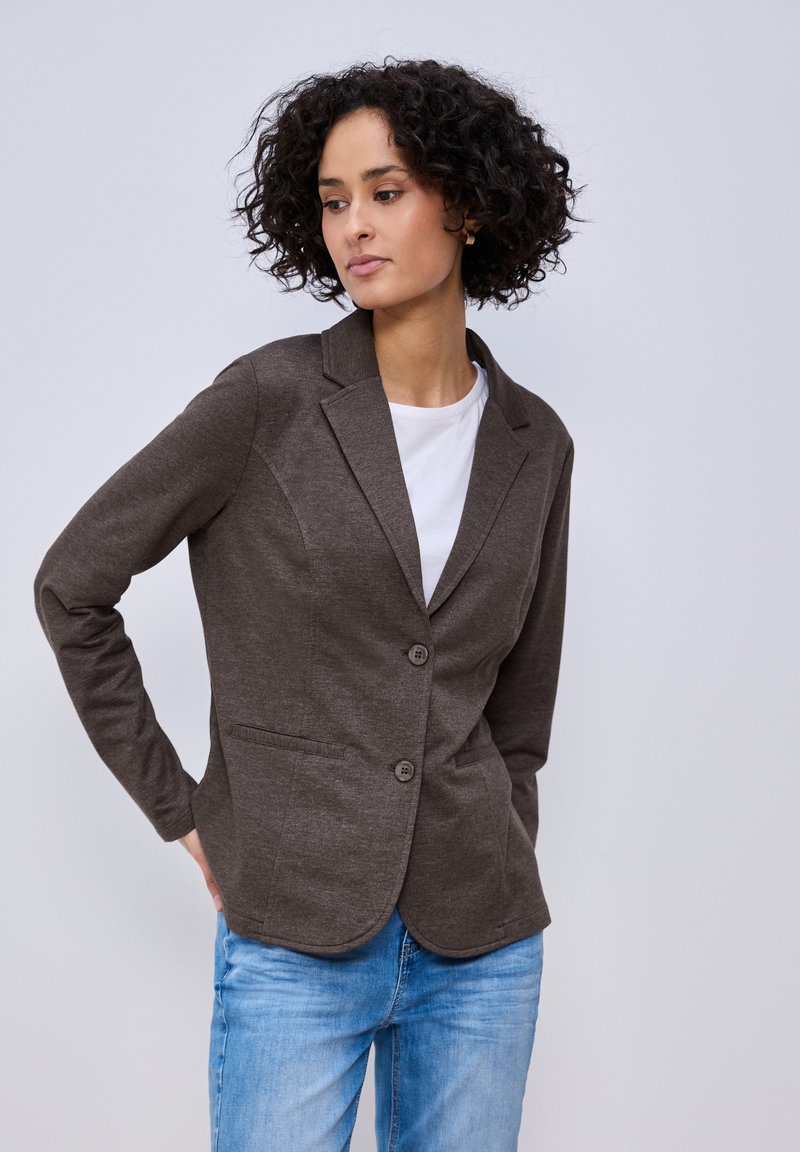 Femme aux cheveux bouclés portant un blazer marron à deux boutons sur une chemise blanche et un jean bleu clair, regardant sur le côté avec les mains derrière le dos.