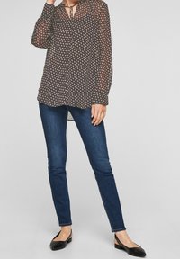 Blusa trasparente scura con un motivo geometrico e scollatura a fiocco, abbinata a jeans attillati blu scuro e scarpe basse nere.