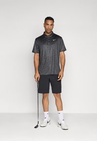 Černé golfové polo tričko s vzorem a límečkem, v kombinaci s tmavými šortkami. Model stojí a drží golfovou hůl, má na sobě bílé tenisky a ponožky.