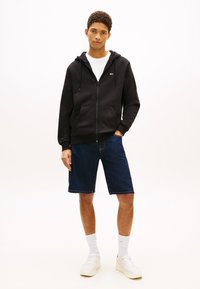 Jauns vīrietis stāv ikdienas apģērbā ar melnu rāvējslēdzēja kapuci, baltu t-kreklu, tumši ziliem džinsa šortiem, baltām sporta kurpēm un baltām zeķēm līdz puslīdzenim.