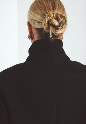 Blond włosy upięte w niski kok, noszone z srebrnymi kolczykami w kształcie kół i czarnym płaszczem z wysokim kołnierzem, widok od tyłu na tle jednolitego tła.