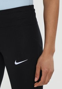 Gros plan sur une personne portant des leggings noirs Nike avec un logo Nike blanc visible et une main détendue reposant sur la cuisse.