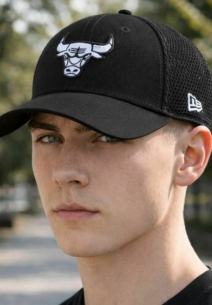 Junger Mann mit heller Haut, der eine schwarze Kappe mit einem Stier-Logo trägt, leicht nach links blickend mit neutralem Gesichtsausdruck im Freien.