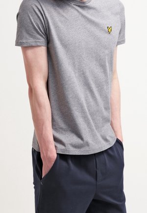 Mężczyzna w krótkim szarym t-shircie z żółtym logo na piersi i granatowych spodniach, z rękami w kieszeniach.
