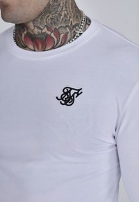 Fehér, hosszú ujjú pamut ing, bal mellen fekete hímzett logóval. Sima textúrájú, kerek nyakkivágású.